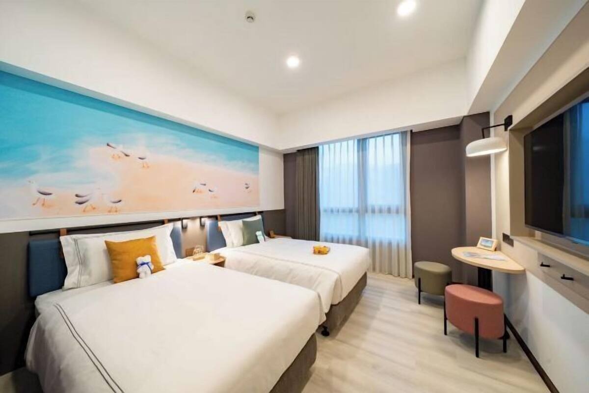 A hotel room with two beds, a small table, a chair, a large window, and a painting on the wall.