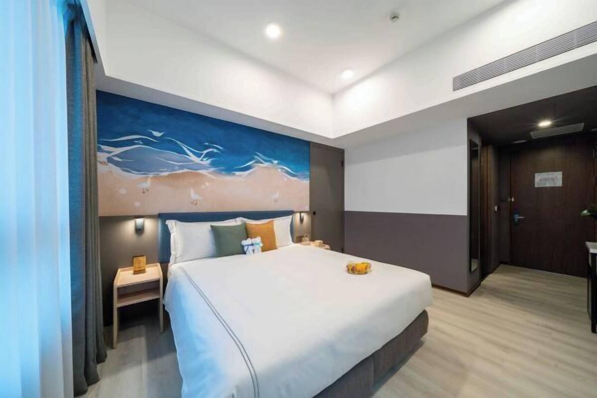 A hotel room with a large bed, a nightstand, a wall-mounted lamp, and a painting of waves.