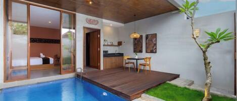 A modern indoor pool area with a wooden deck, a dining table, and a small garden with a tree.