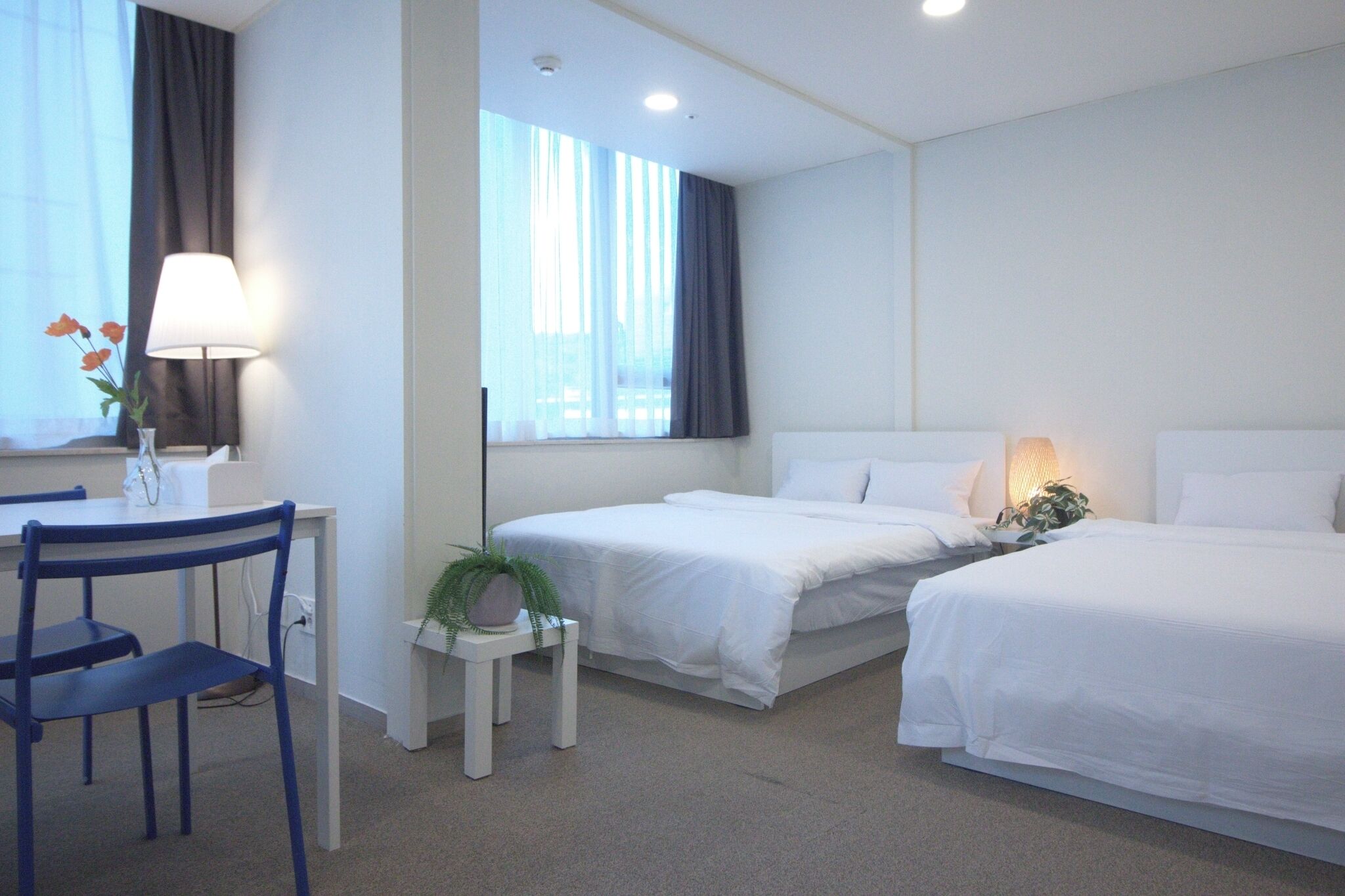 Quarto de hotel com duas camas, uma escrivaninha com cadeira, uma mesinha com uma planta e uma janela com cortinas.