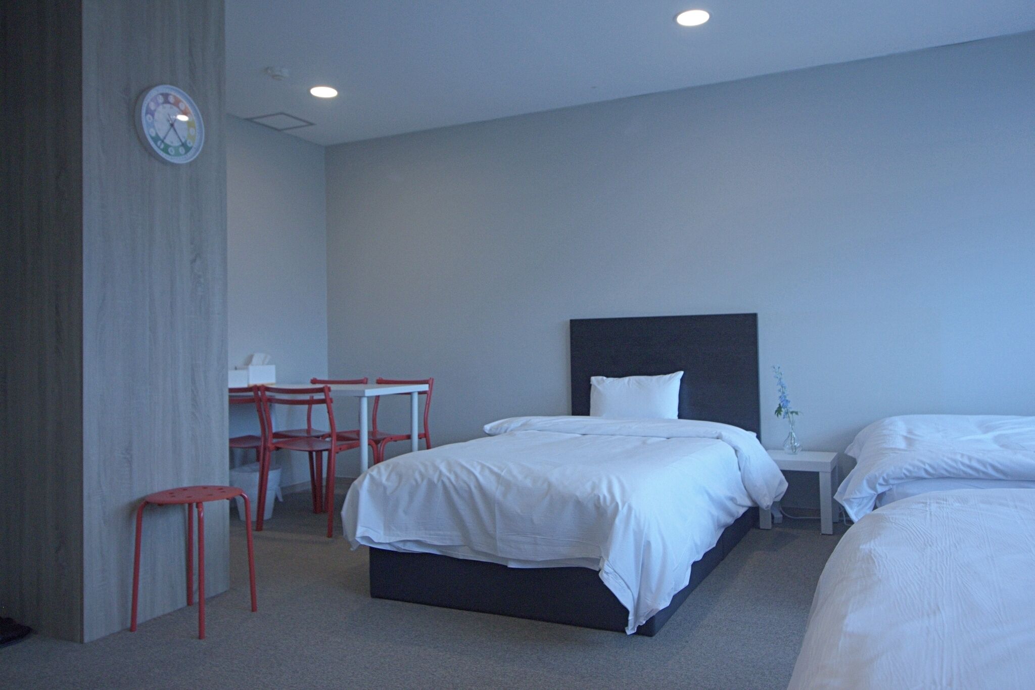 A hotel room with a bed, bedside table, chair, and a clock on the wall.