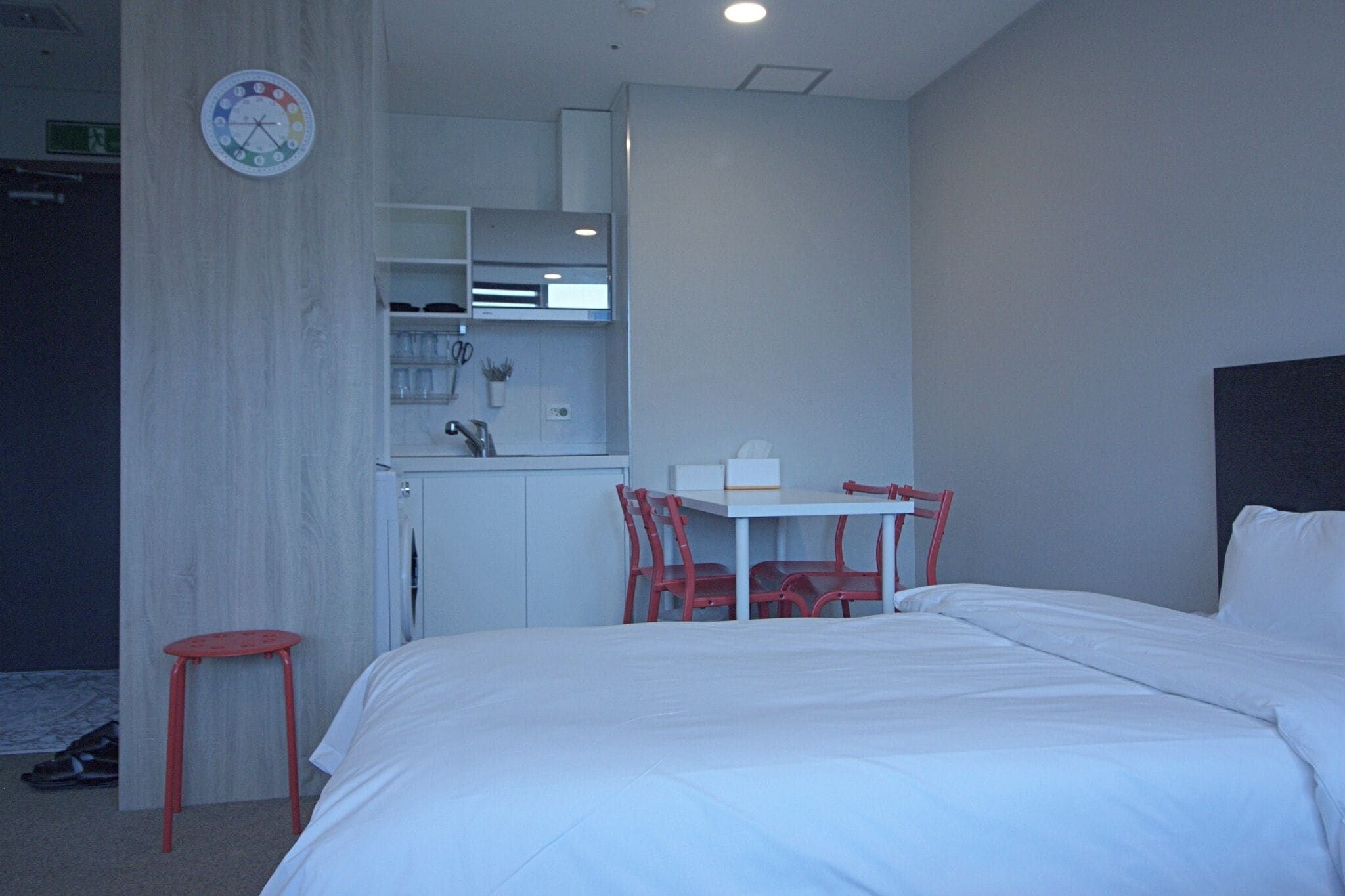 A hotel room with a bed, a small table with chairs, a kitchenette, and a clock on the wall.