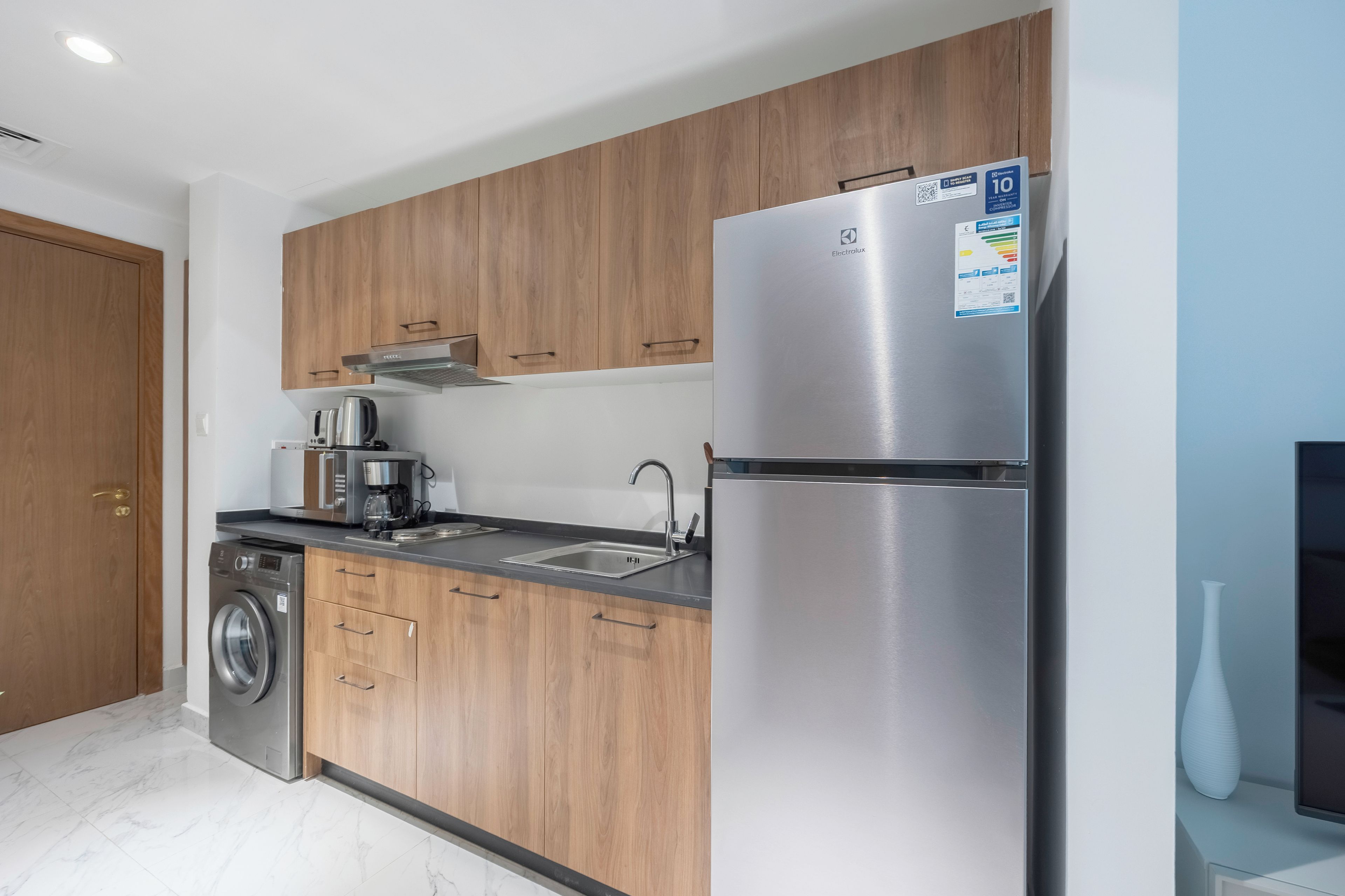 A modern kitchen with wooden cabinets, a stainless steel refrigerator, a coffee maker, and a washing machine.