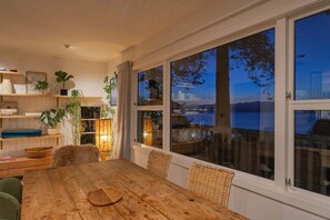 Uma área de refeições com mesa de madeira e cadeiras de vime, grandes janelas com vista para um corpo d'água, e prateleiras com plantas e livros.