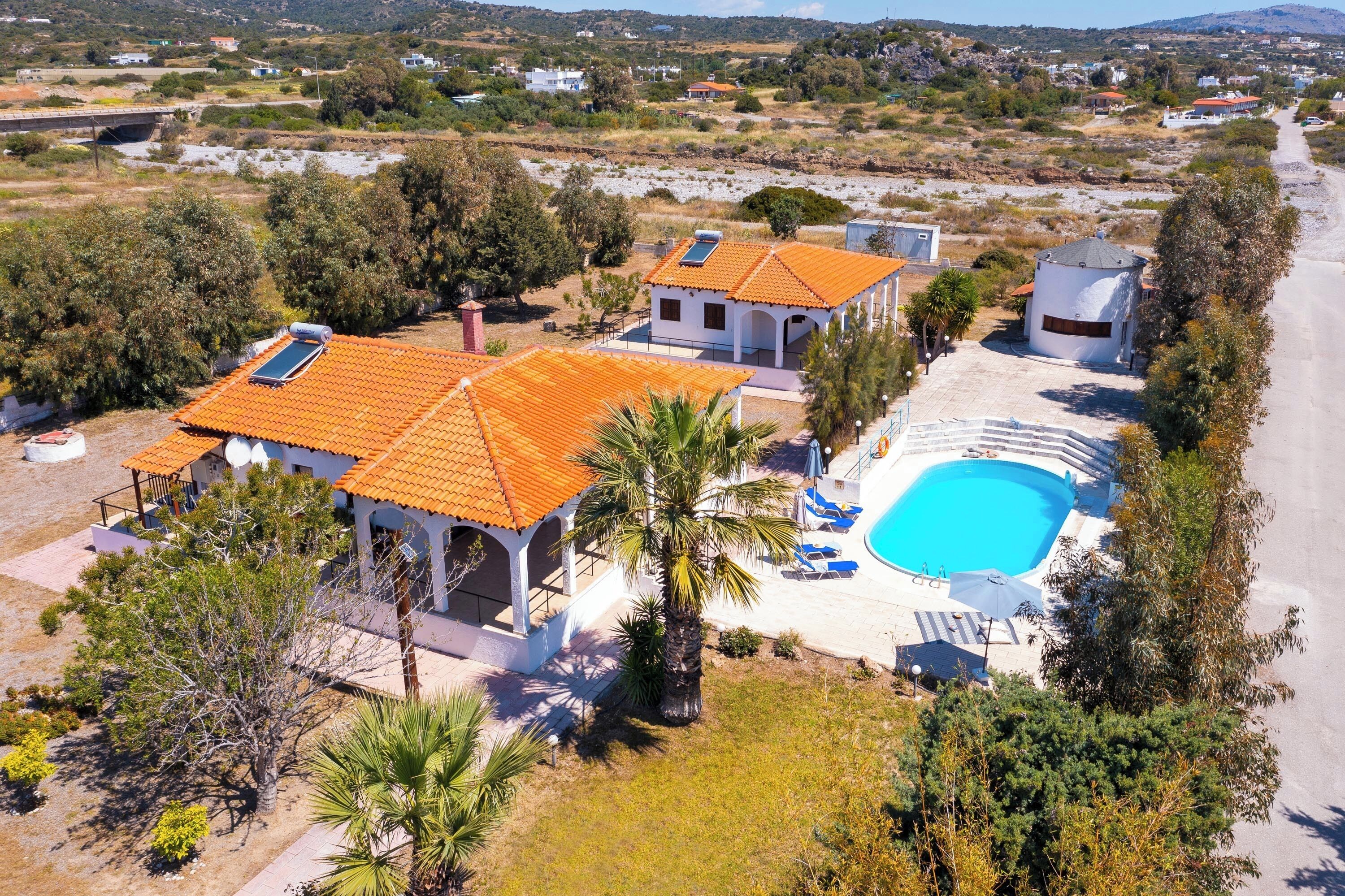 A residential property with a swimming pool, multiple houses with orange tiled roofs, and surrounding greenery.
