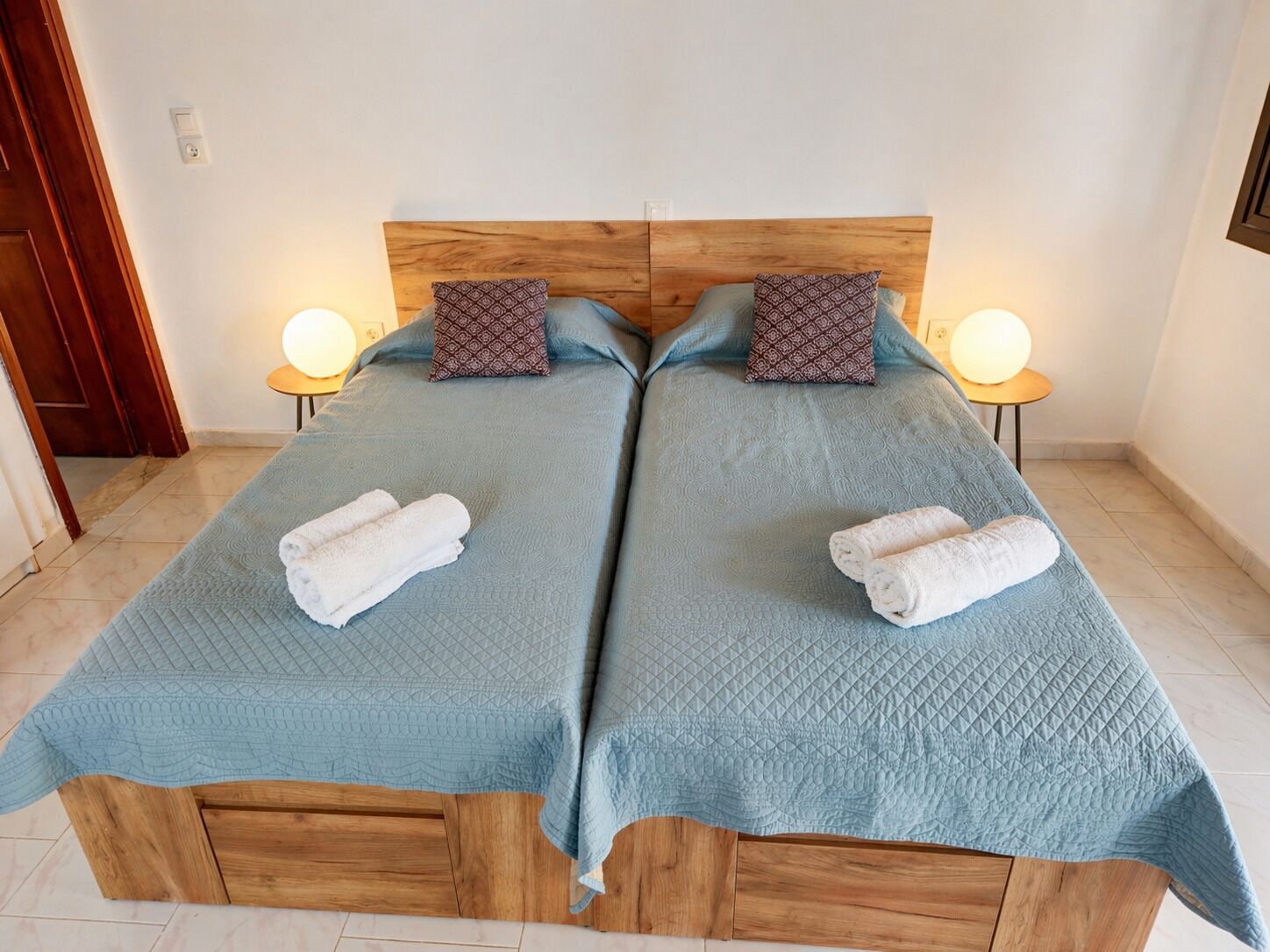 A double bed with a wooden headboard, two pillows, two bedside lamps, and two rolled towels.
