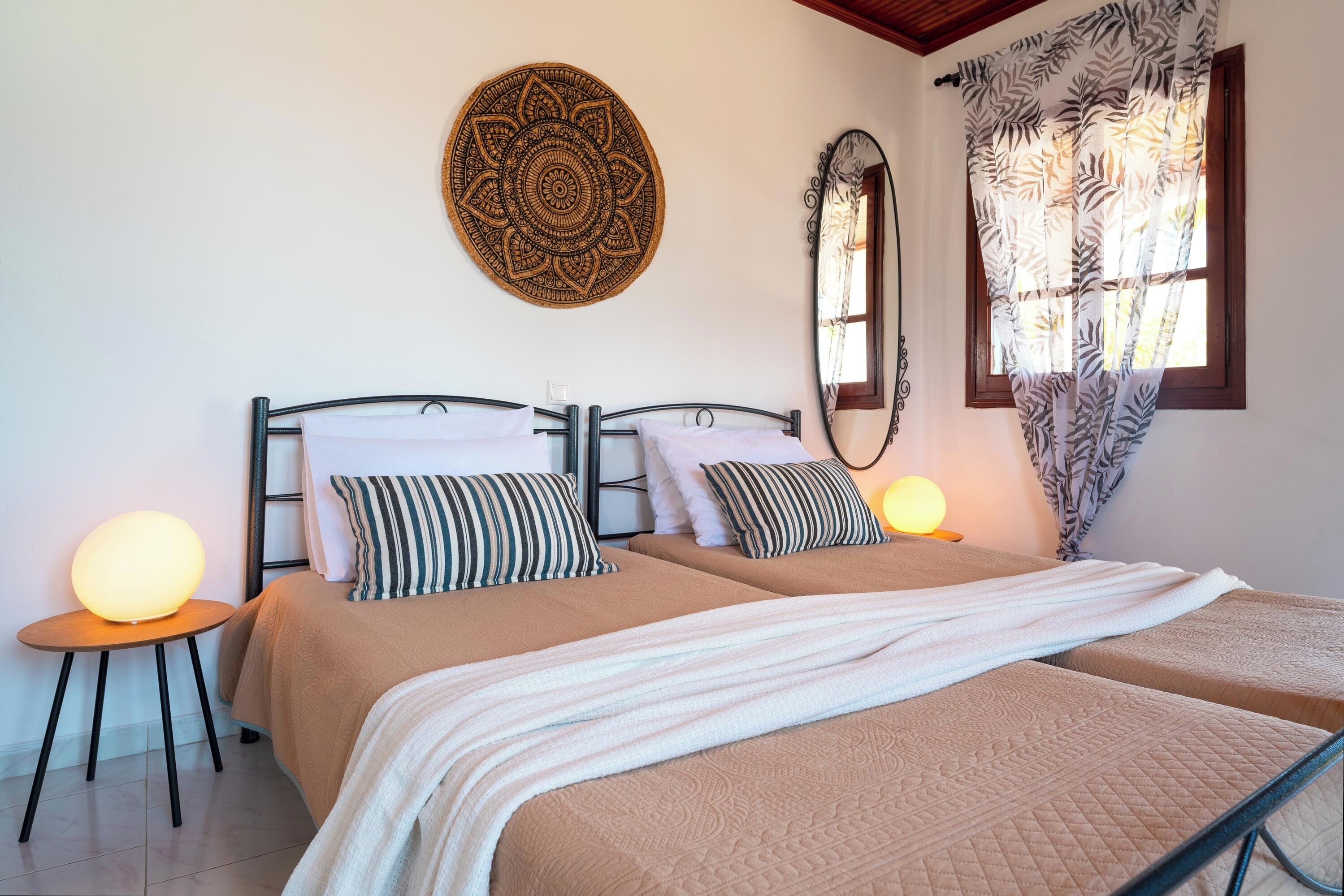 Uma cama de casal com cobertura acolchoada bege, duas mesas de cabeceira com luminárias, um espelho fixado na parede e um ornamento circular decorativo na parede.