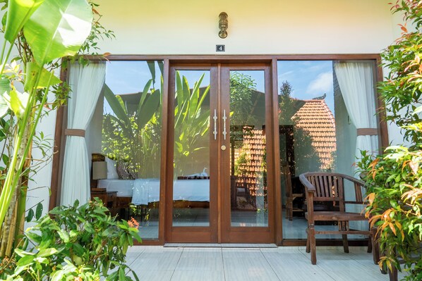 A sliding glass door entrance to a room with a bed, a chair, and a hanging light fixture. - Dream Breeze Village (Pecatu)