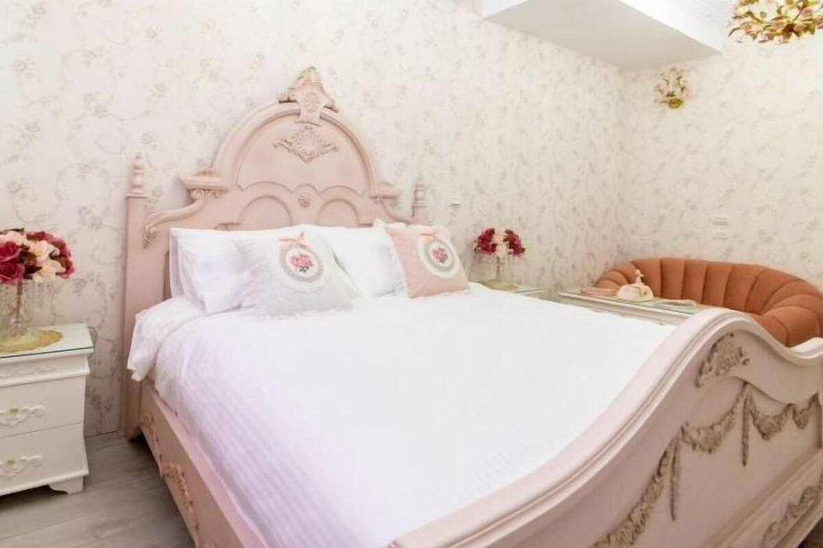 A bedroom with a pink bed, white bedding, and floral wallpaper.