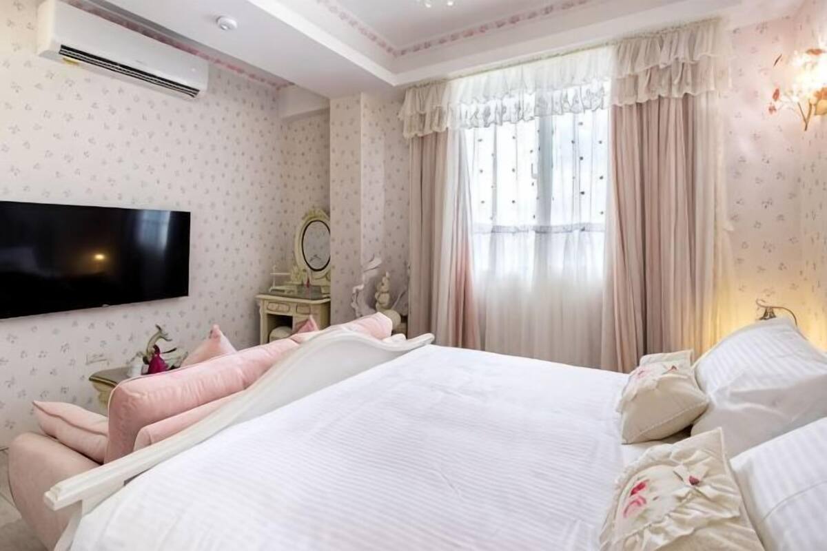 A bedroom with a large bed, a television, a vanity with a mirror, and a window with curtains.