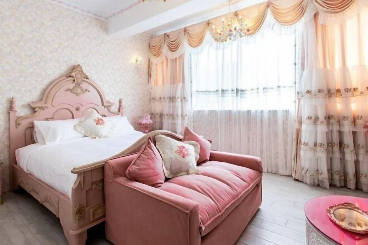 A bedroom with a pink bed, a matching pink sofa, and a chandelier.
