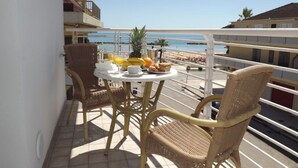 Ein Balkon mit Korbstühlen und einem Tisch, gedeckt mit Frühstücksleckereien, mit Blick auf einen Strand und Gebäude.
