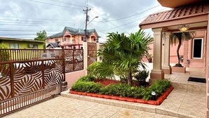 Una entrada protegida por un cerramiento decorativo, un jardín bien cuidado con diversas plantas, y un edificio con tejado de baldosas y un balcón.