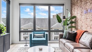 Een moderne woonkamer met een groot raam, een blauwe fauteuil, een leren bank en een bakstenen muur.