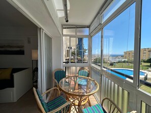 A balcony with wicker chairs and a glass table, overlooking a swimming pool and buildings. - Apartment with a large terrace and ocean view (Torremolinos)