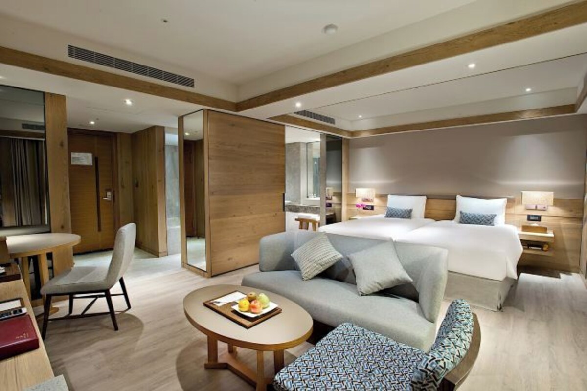 A modern hotel room with a bed, sofa, armchair, and a small table with fruit.