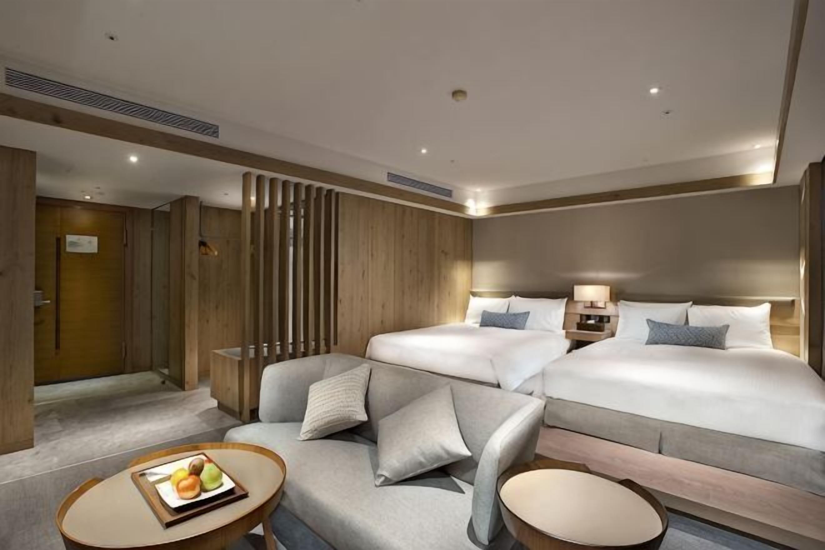 A hotel room with a double bed, a sofa, a small table with fruit, and a decorative wall panel.