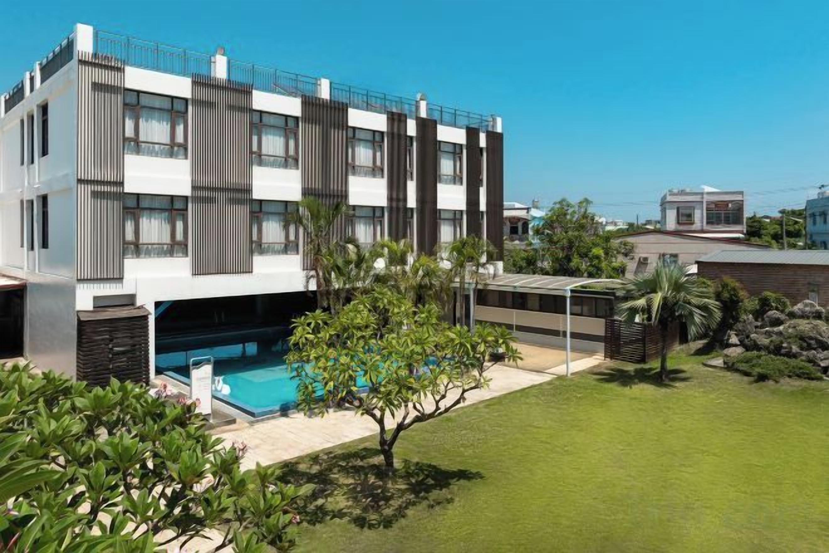 A modern multi-story building with a swimming pool, surrounded by a well-maintained lawn and some trees.