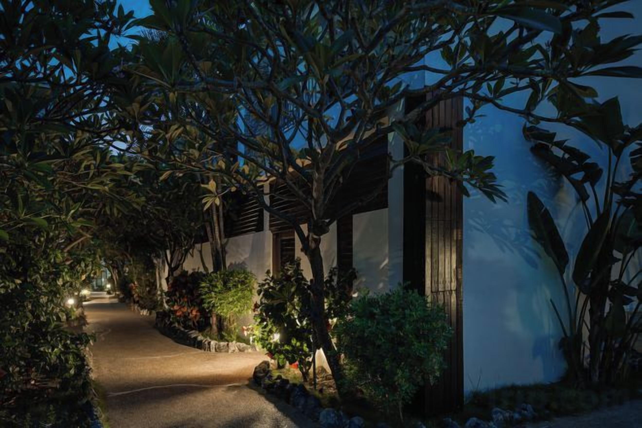 Un camino conduce a un edificio con acceso cubierto, rodeado de vegetación exuberante e iluminado por luz ambiental.