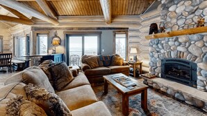 A cozy log cabin interior with a stone fireplace, wooden ceiling beams, and a large window overlooking a snowy landscape. - Alpine Ridge on Whitefish Mountain Sleeps 12 (Whitefish)