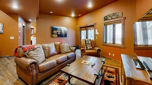 A cozy living room with a leather sofa, a wooden coffee table, and a chair with a floral cushion. A painting hangs on the wall, and there are windows with curtains. - Alpine Ridge on Whitefish Mountain Sleeps 12 (Whitefish)