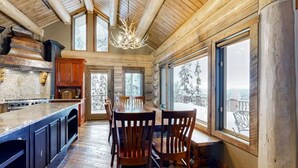 A log cabin interior with a dining area, kitchen, and large windows offering a view of snow-covered trees. - Alpine Ridge on Whitefish Mountain Sleeps 12 (Whitefish)
