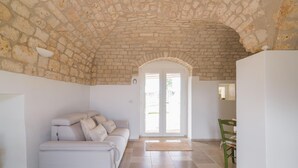Ένα δωμάτιο με πέτρινη οροφή, έναν λευκό καναπέ, ένα τραπέζι φαγητού με καρέκλες και μια μεγάλη αψιδωτή είσοδο.