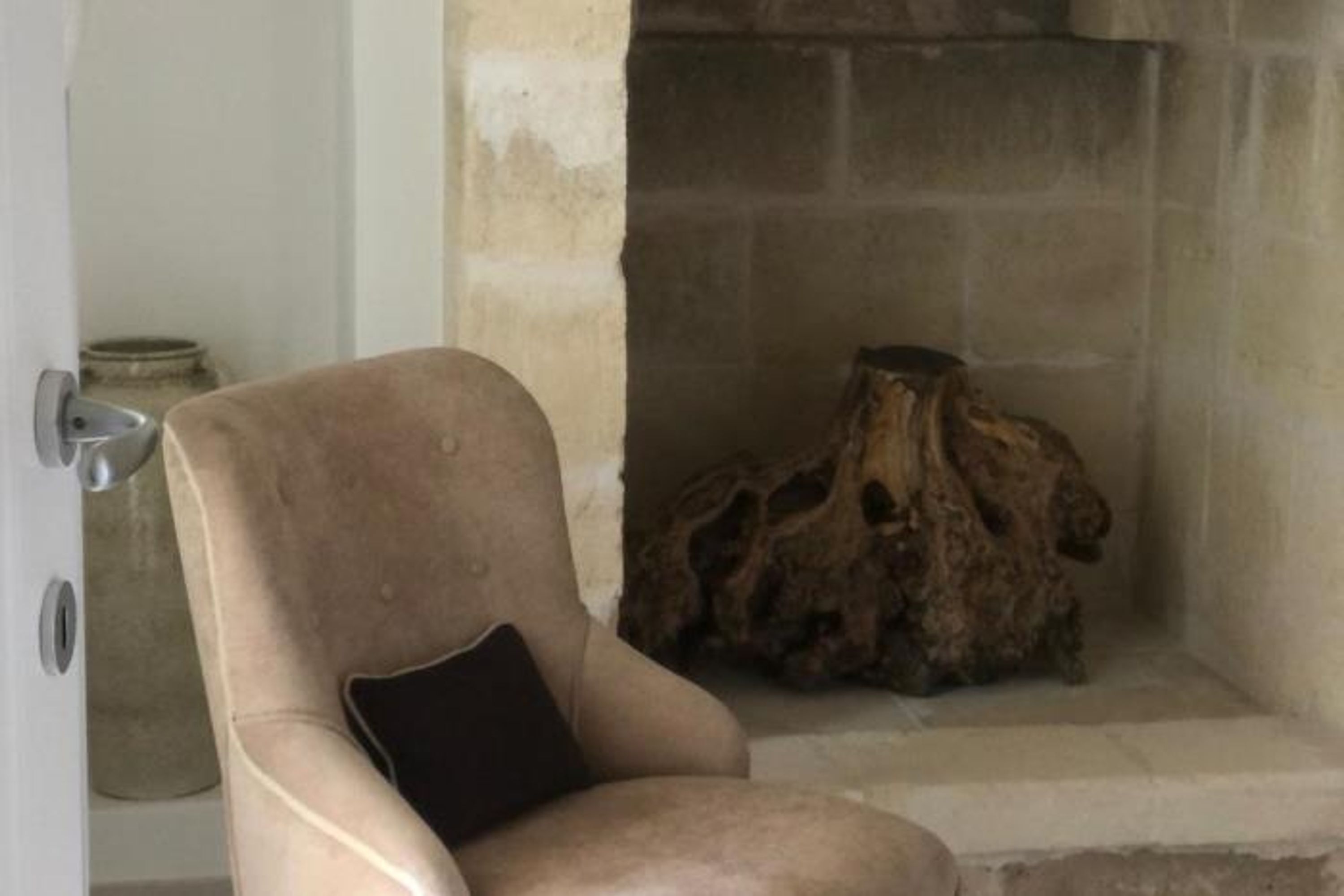 A beige armchair with a black cushion in a room with a stone fireplace and a rustic wooden log.