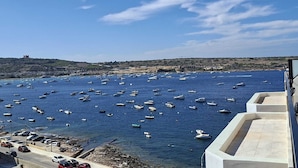Vista costera con numerosos barcos anclados en el agua, una carretera con coches aparcados y una colina distante con una estructura en la cima.