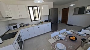 Una cocina moderna con armarios blancos, un refrigerador de acero inoxidable y un comedor para cuatro personas con una mesa, sillas y un frutero.