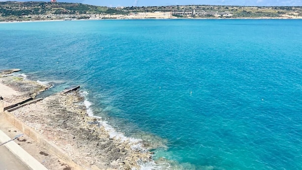 Vista costera con agua azul cristalina, una costa rocosa y edificios a lo lejos.