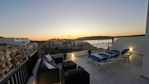 Una terraza en la azotea con sillones de descanso y un sofá, con vistas a un paisaje urbano y una masa de agua al atardecer.