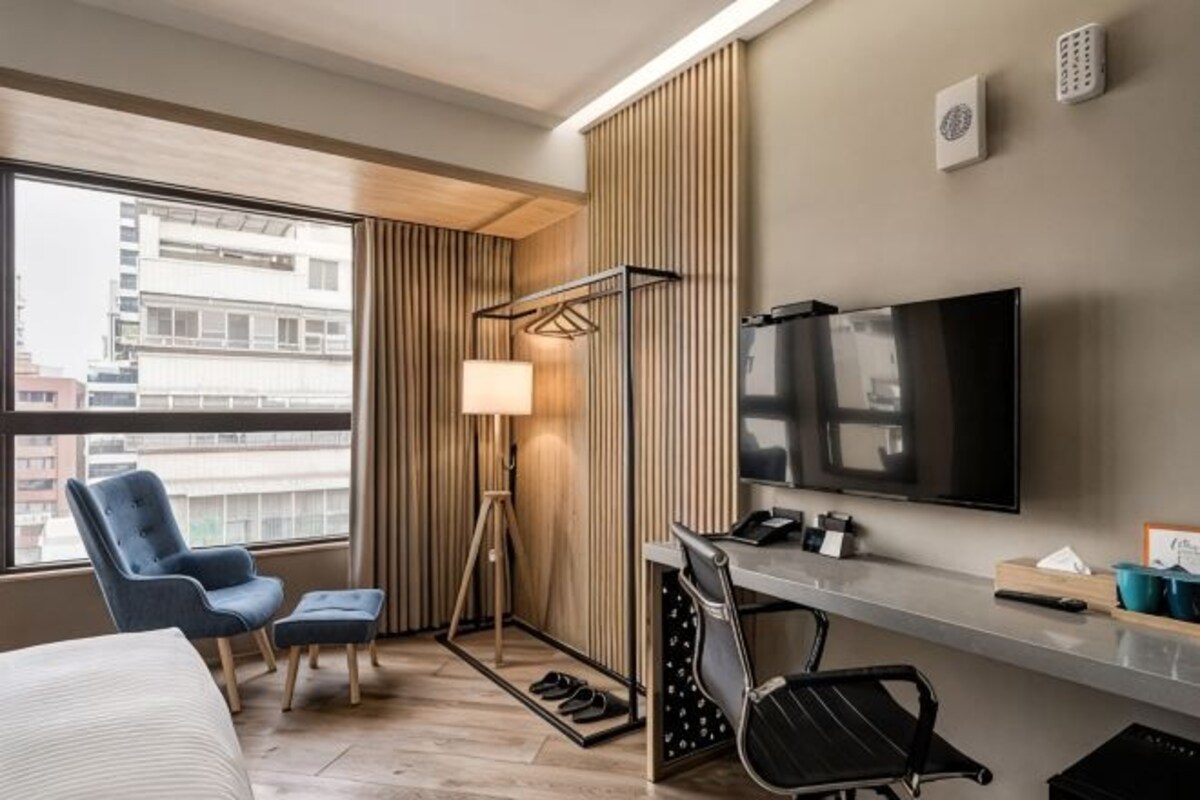Habitación de hotel con cama, escritorio con televisor, silla, lámpara y vista a la ciudad.