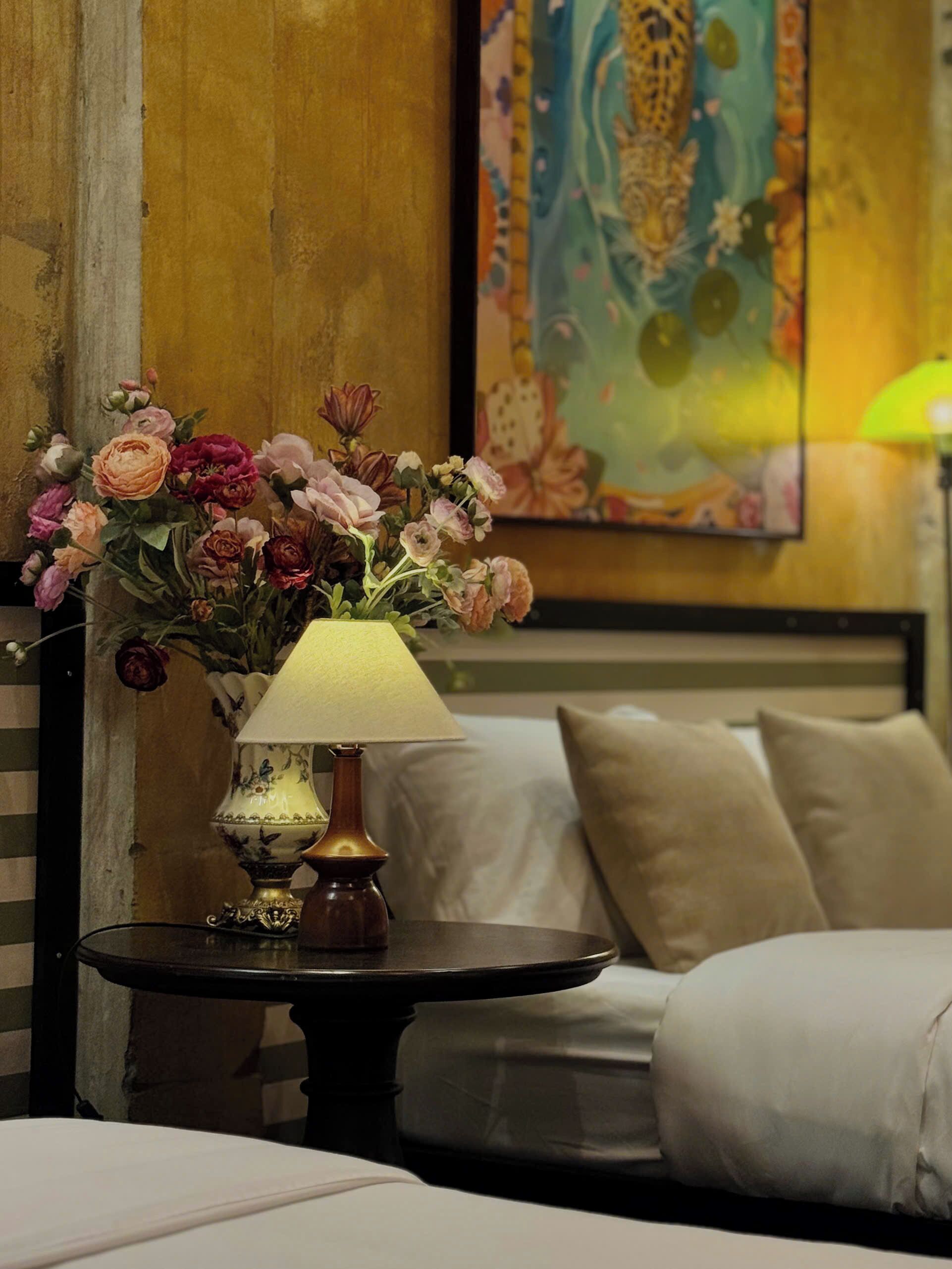 A room with a bed, a small table with a lamp, and a painting on the wall.