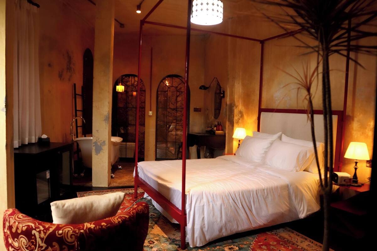 A four-poster bed with white linens, a red chair with a patterned cushion, a wooden floor, and a chandelier.