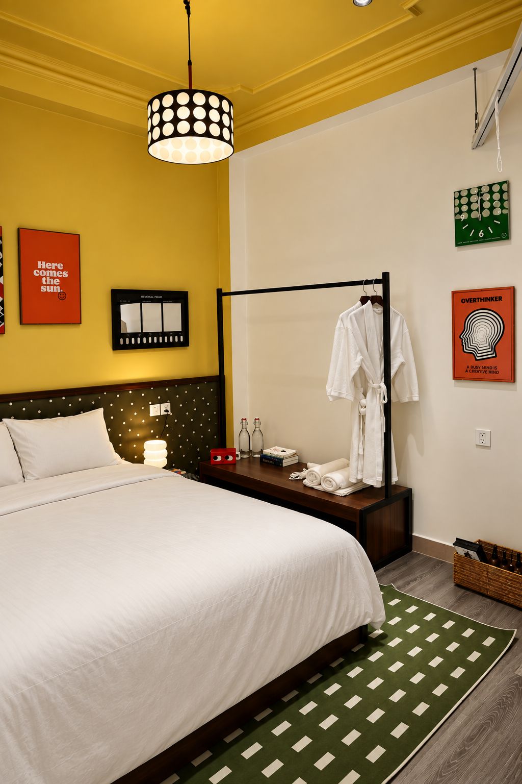 A hotel room with a bed, a bedside table, a nightstand, a wall-mounted clock, a towel rack, and a green patterned rug.