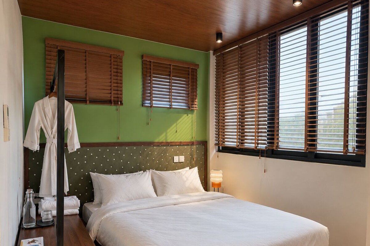 Un dormitorio moderno con cabecera de madera, una cama grande, una mesita de noche, una bata y una ventana con persianas.
