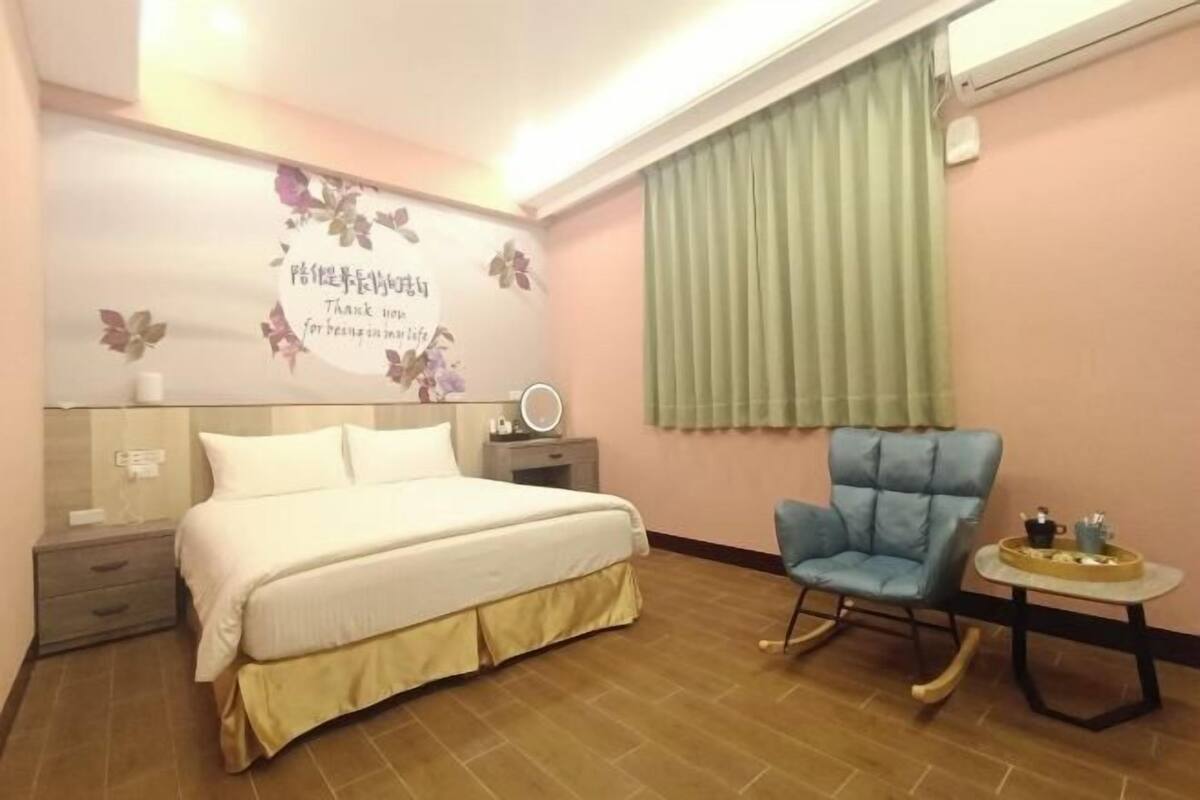 A bedroom with a bed, a chair, a bedside table, a mirror, and a wall with a floral design.