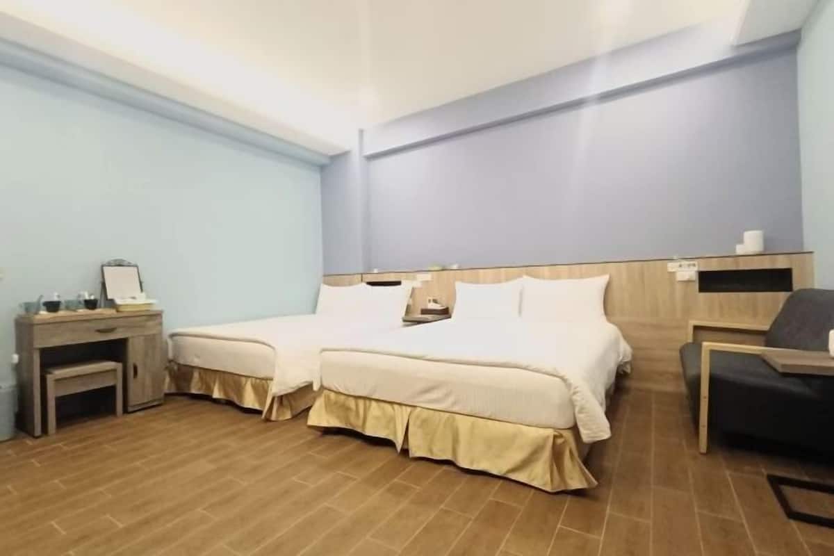 A hotel room with a double bed, a desk with a chair, a small table, and a shelf with toiletries.