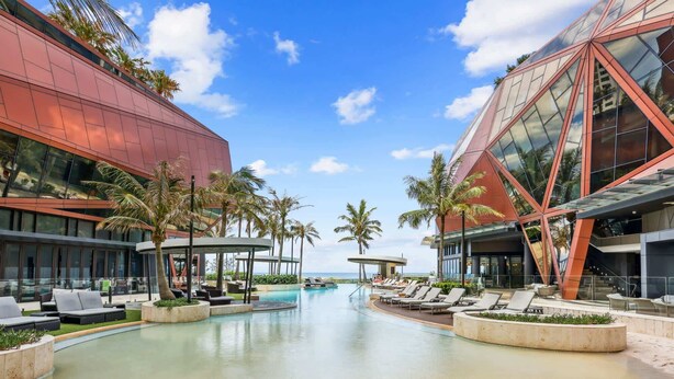 A modern hotel with a large pool, palm trees, and a clear blue sky. - Luxury Beachfront 1-Bed Apartment with Pool & Gym (Surfers Paradise)