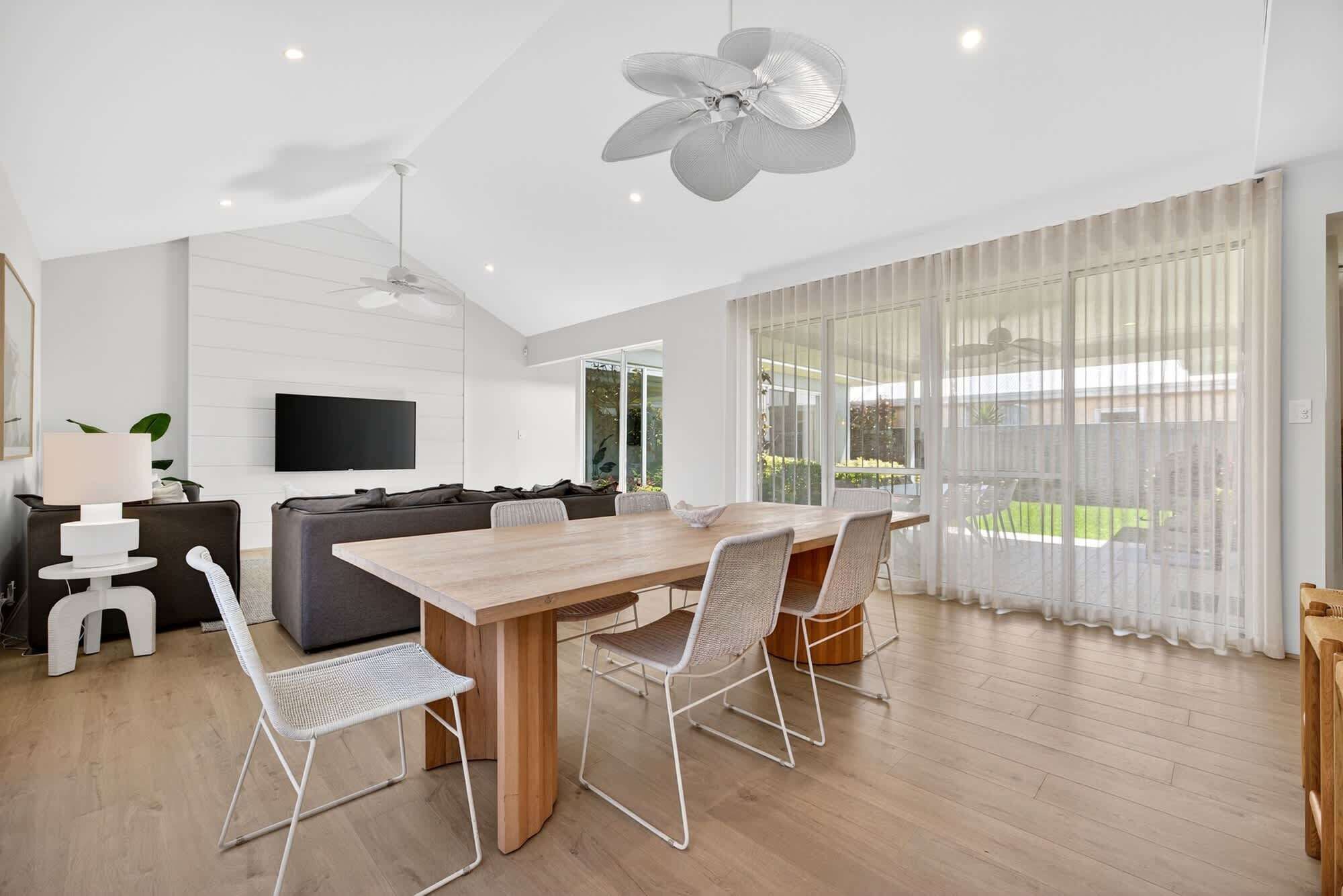 A modern living room with a wooden dining table, white chairs, a grey sofa, a flat-screen TV, and a ceiling fan.