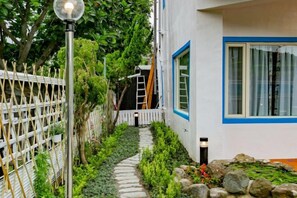 Un étroit sentier de pierre mène à un bâtiment blanc orné de bordures bleues et doté d’une fenêtre avec des rideaux.
