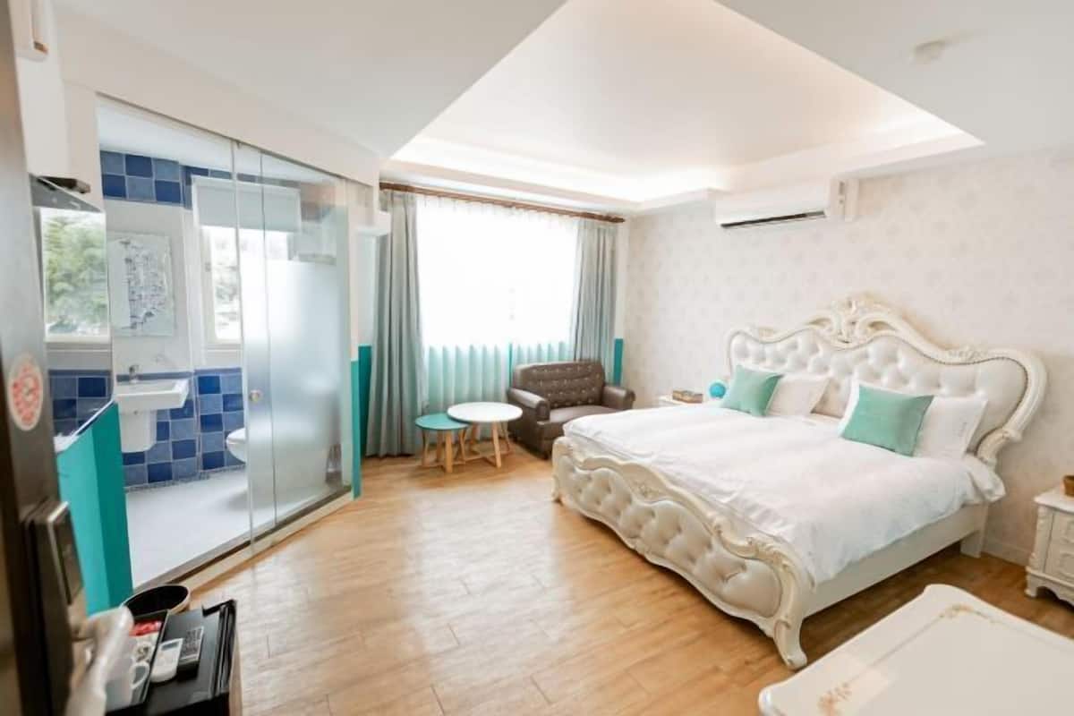 Un dormitorio con una cama grande, una silla, una mesita pequeña y una puerta corrediza de vidrio que da al baño.
