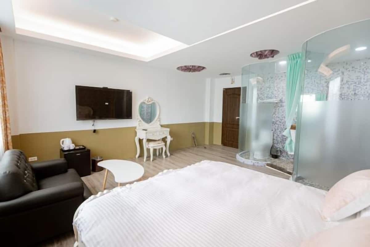 Habitación de hotel con cama, televisión, escritorio, silla y cabina de ducha con paredes de vidrio.