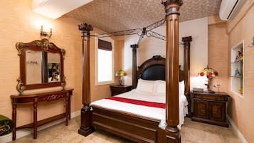 A four-poster bed with a dark headboard, a wooden console table with a mirror, and a small bedside table with a lamp and flowers.