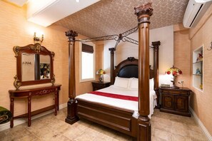 Ein Bett mit Baldachin, ein dunkles Kopfteil, ein Holztischchen mit Spiegel und ein Nachttisch mit Lampe und Blumen.