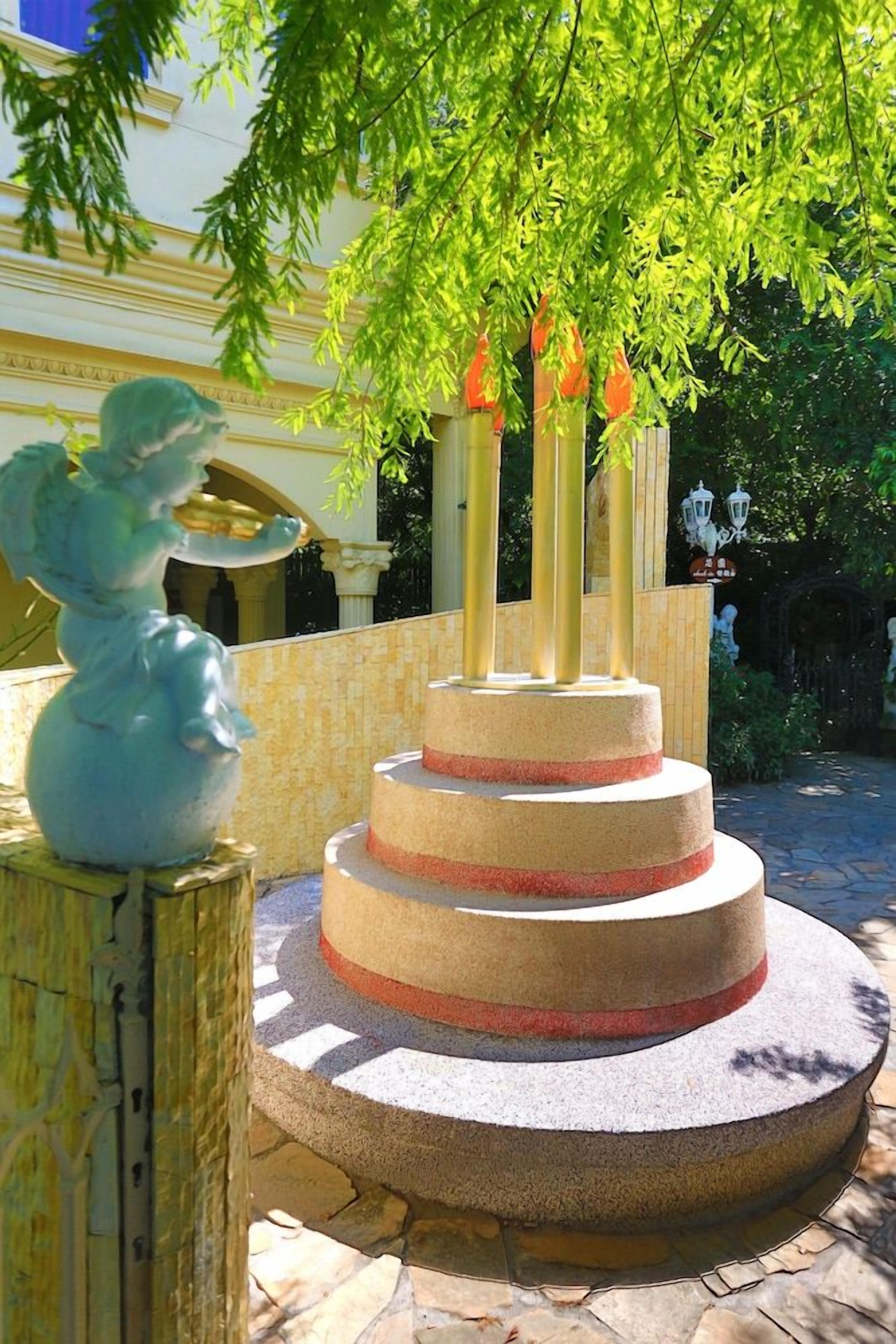 Una fuente de piedra de varios niveles con una estatua de un ángel sobre un pedestal, rodeada de vegetación y un edificio al fondo.