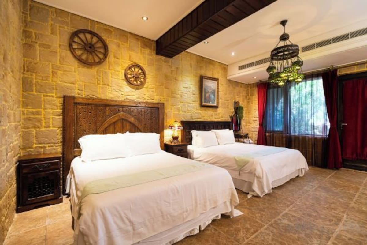 A room with two beds, stone walls, a wooden headboard, a clock, and a chandelier.