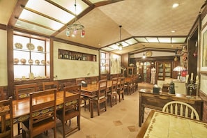 Un grand espace dédié à la salle à manger, avec des tables et des chaises en bois, un bar avec des étagères remplies de bouteilles, et une horloge au mur.