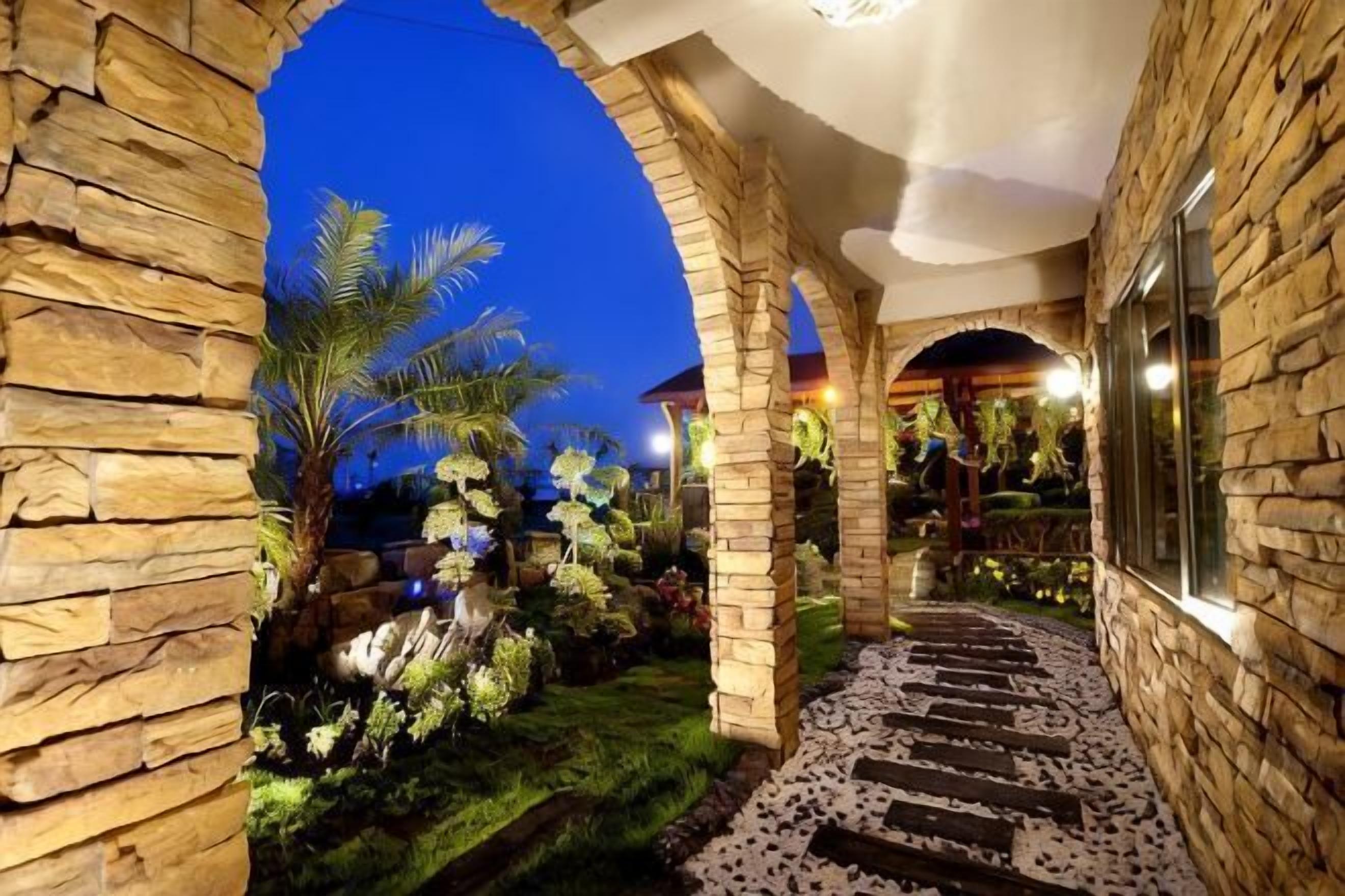A stone archway leading to a garden with a stone pathway and palm trees.